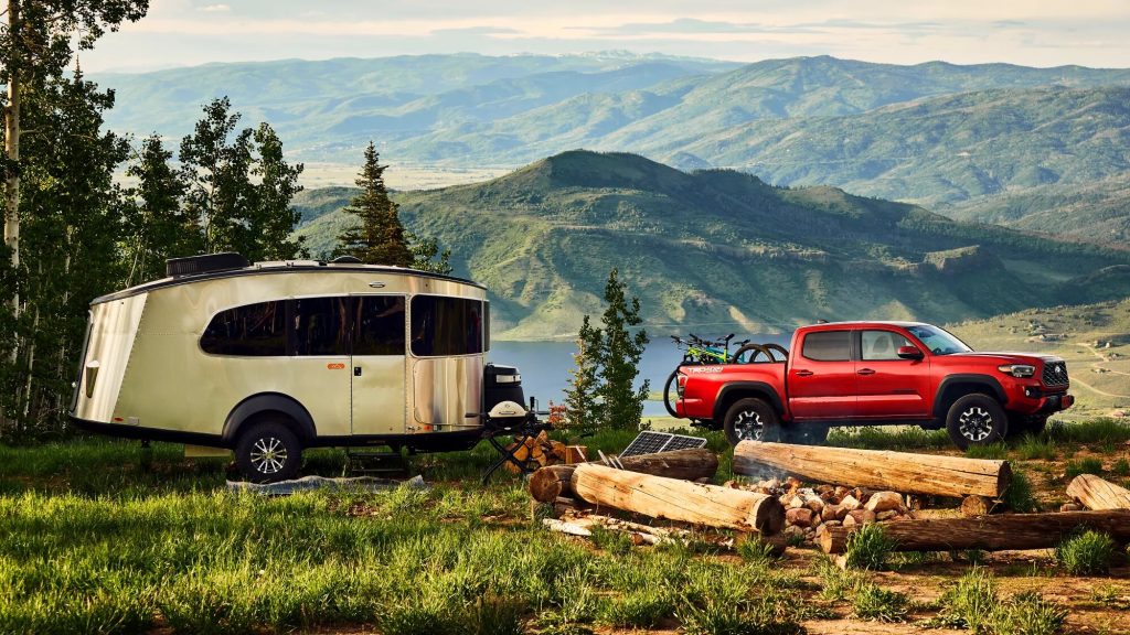 Red 2022 Toyota Tacoma towing a camper on a campground with a beautiful mountain scene in the background.