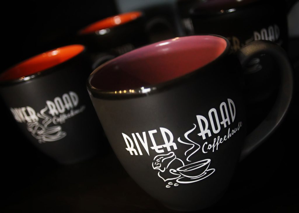 Up-close shot of 3 black mugs with white font spelling 'River Road Coffeehouse'. 