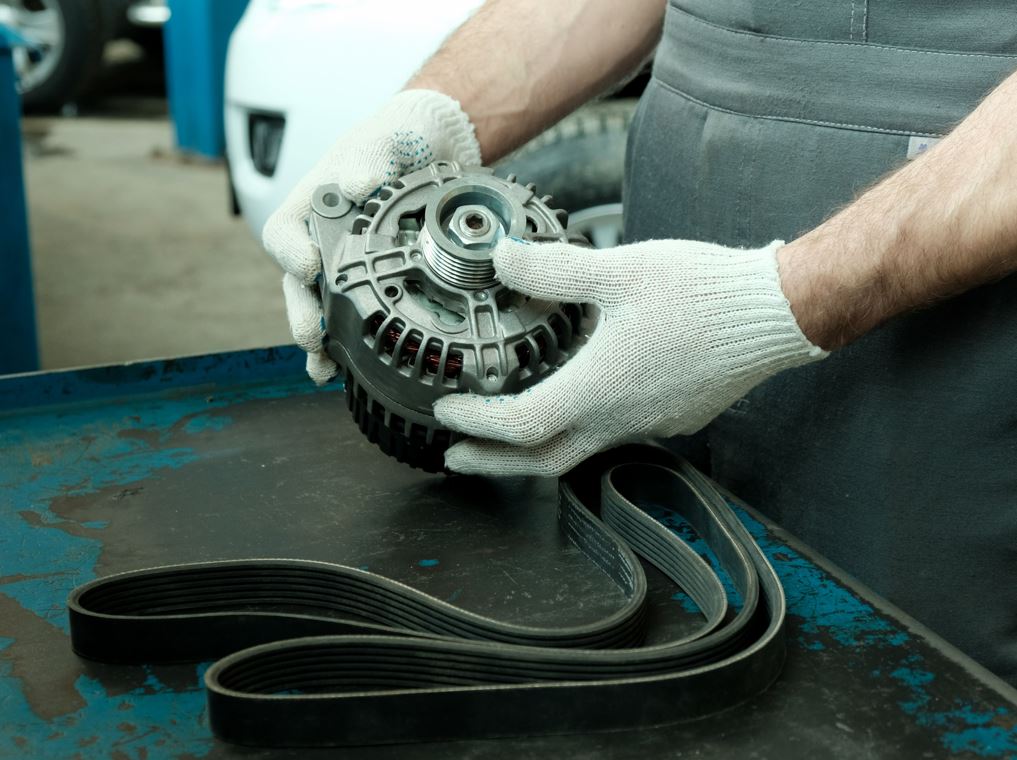 Gloved hands can be seen holding a vehicle alternator.