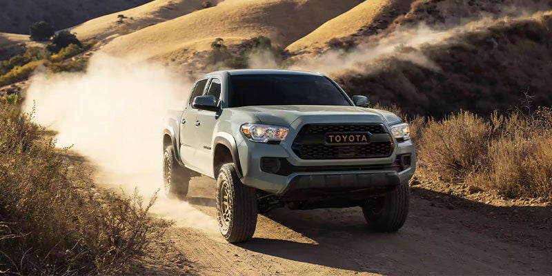 A white 2023 Toyota Tacoma driving towards us in perspective on a dirt road, with dust blowing in the back.