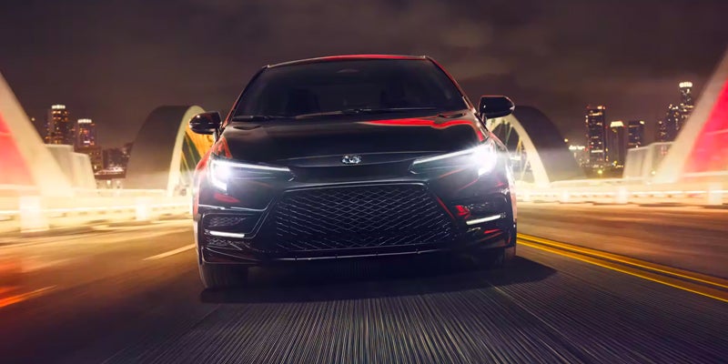 A 2024 Toyota Corolla driving in the night with its headlights blaring. The exterior color of the car is dark, possibly black or dark gray.